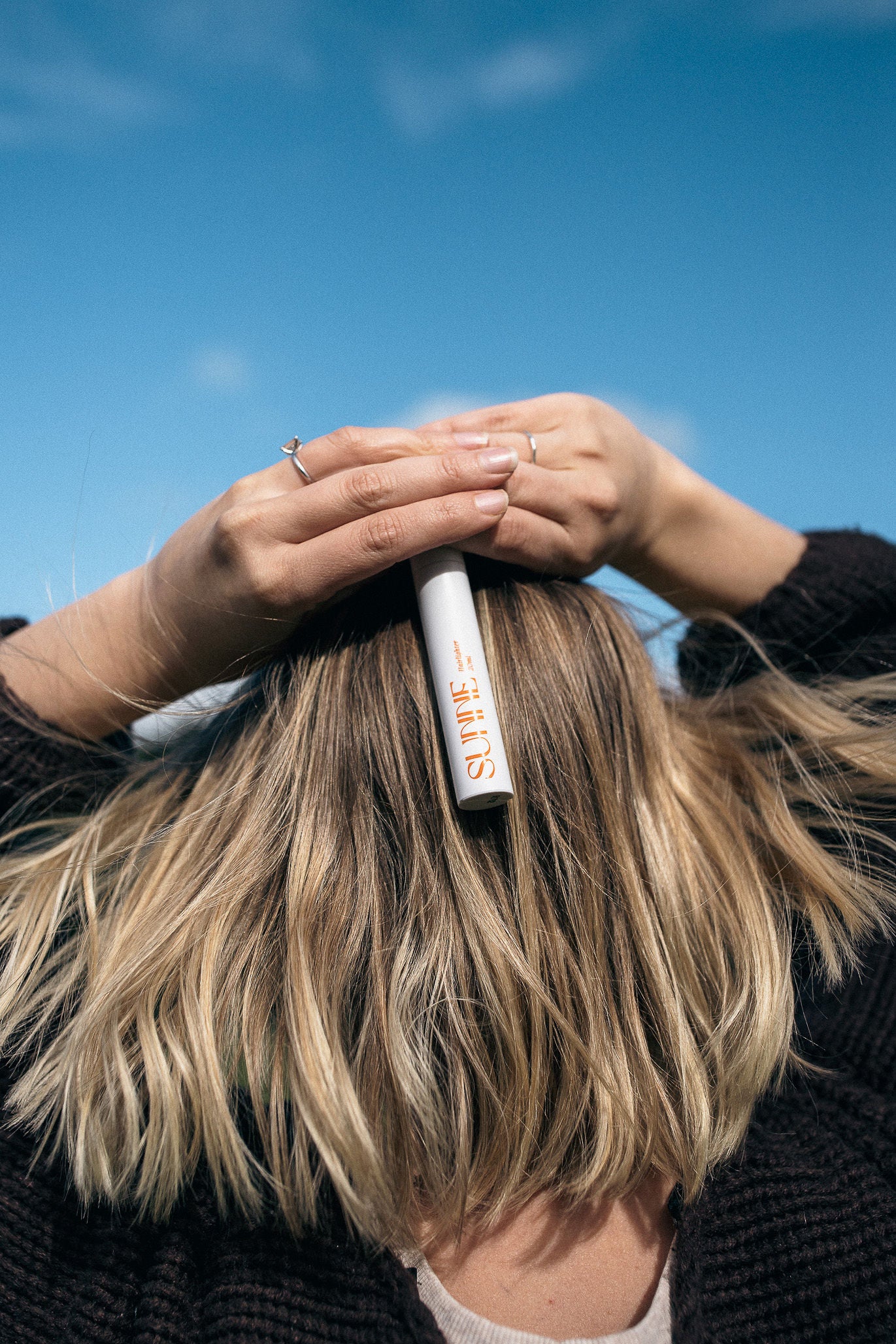 A woman holds the Sunne Hairlighter over her blonde hair under a blue sky, showing how the heat-activated hair lightening spray creates natural, sun-kissed highlights.